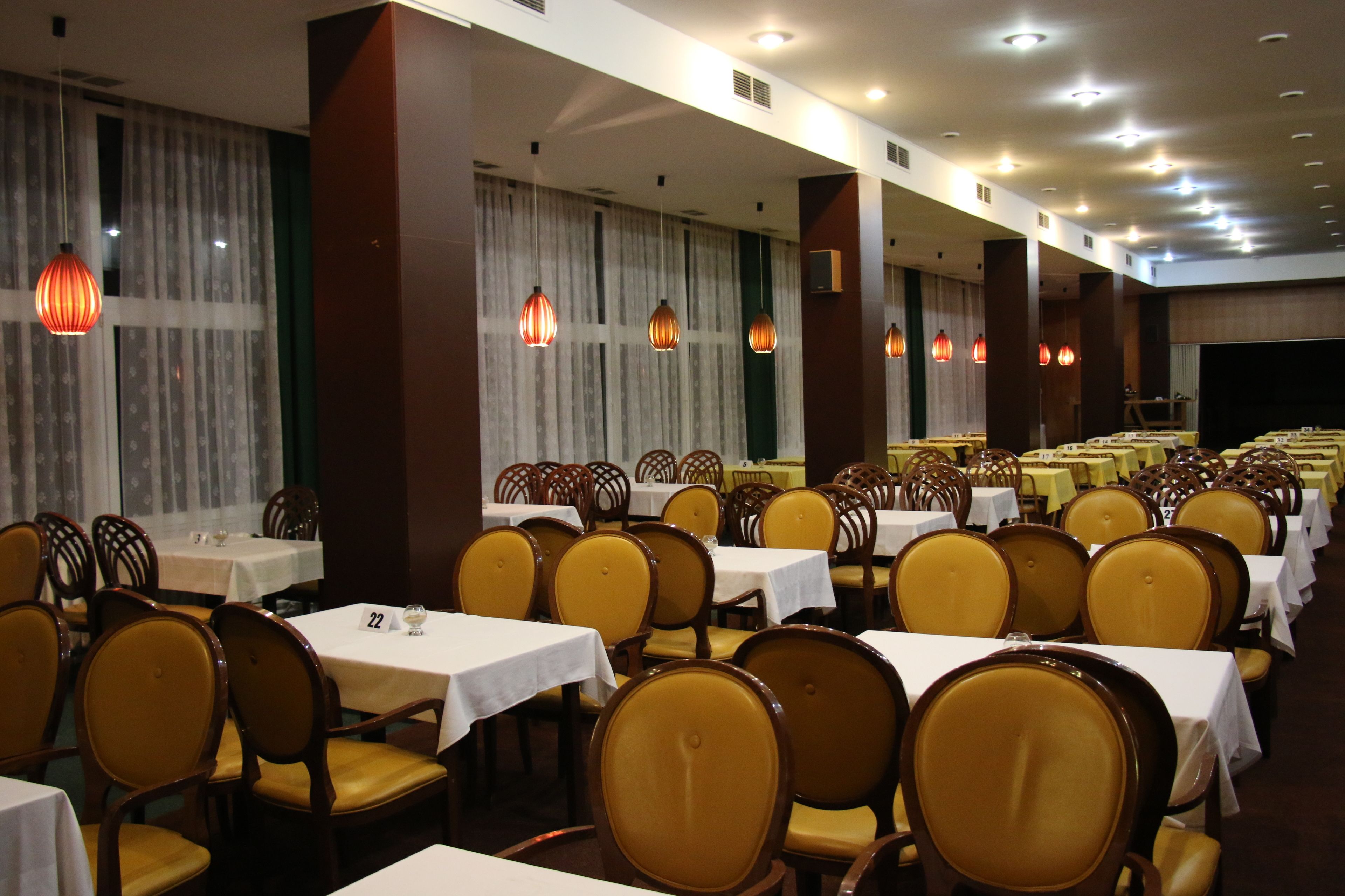 Une salle de banquet avec des rangées de tables et de chaises, de grandes fenêtres ornées de rideaux et des luminaires suspendus.