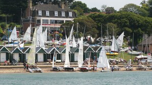 Un port de plaisance où sont amarrés plusieurs voiliers, un bâtiment avec une enseigne et une zone herbeuse où se trouvent des personnes.