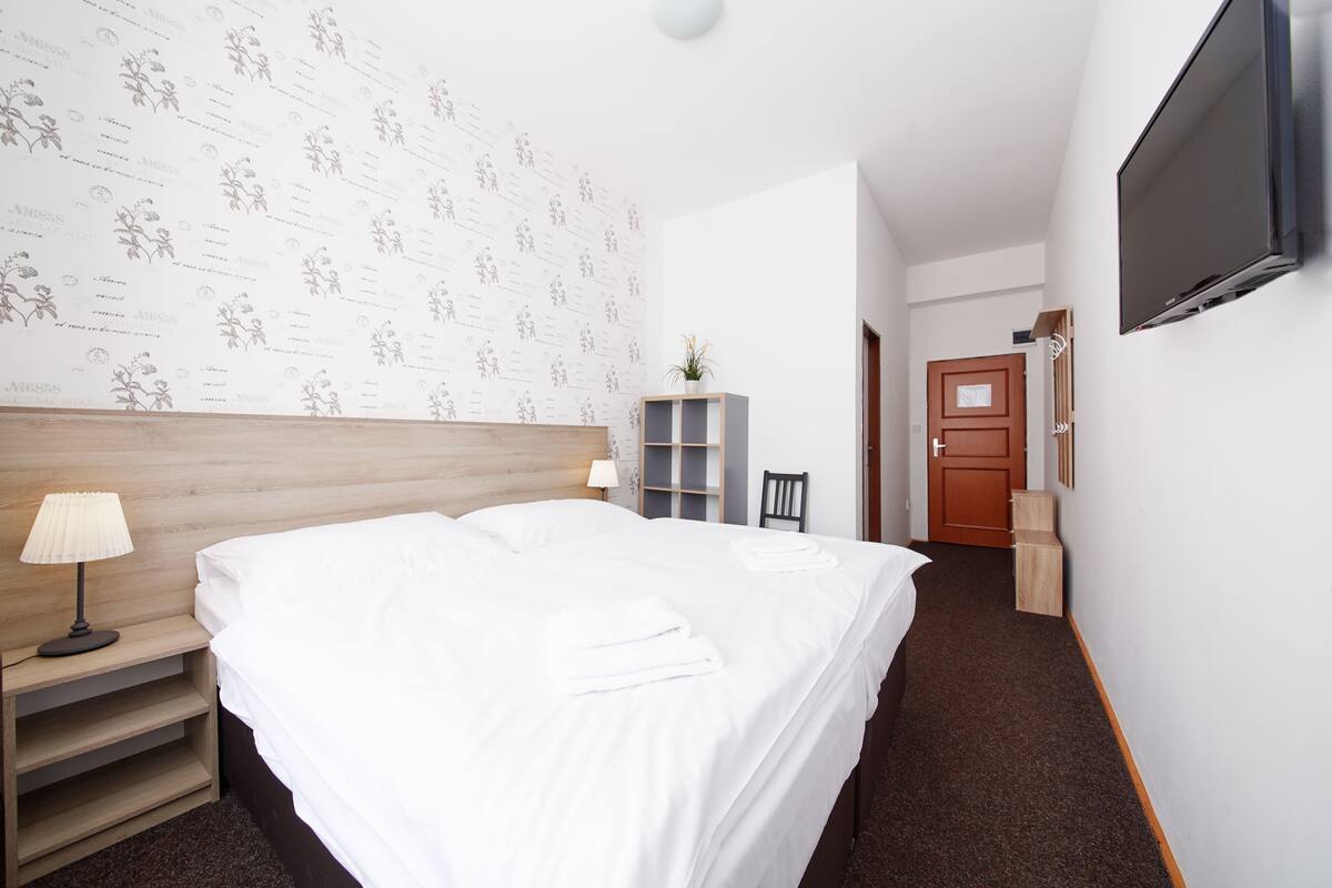 Une chambre d’hôtel avec une tête de lit en bois, un lit avec du linge de maison blanc, une table de chevet avec une lampe, un téléviseur fixé au mur et une porte donnant sur une autre pièce.