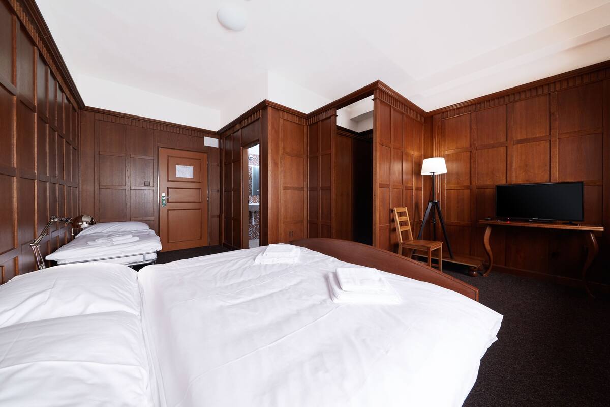 Une chambre d’hôtel avec trois lits, des panneaux de bois au mur, une télévision et un bureau avec une lampe.