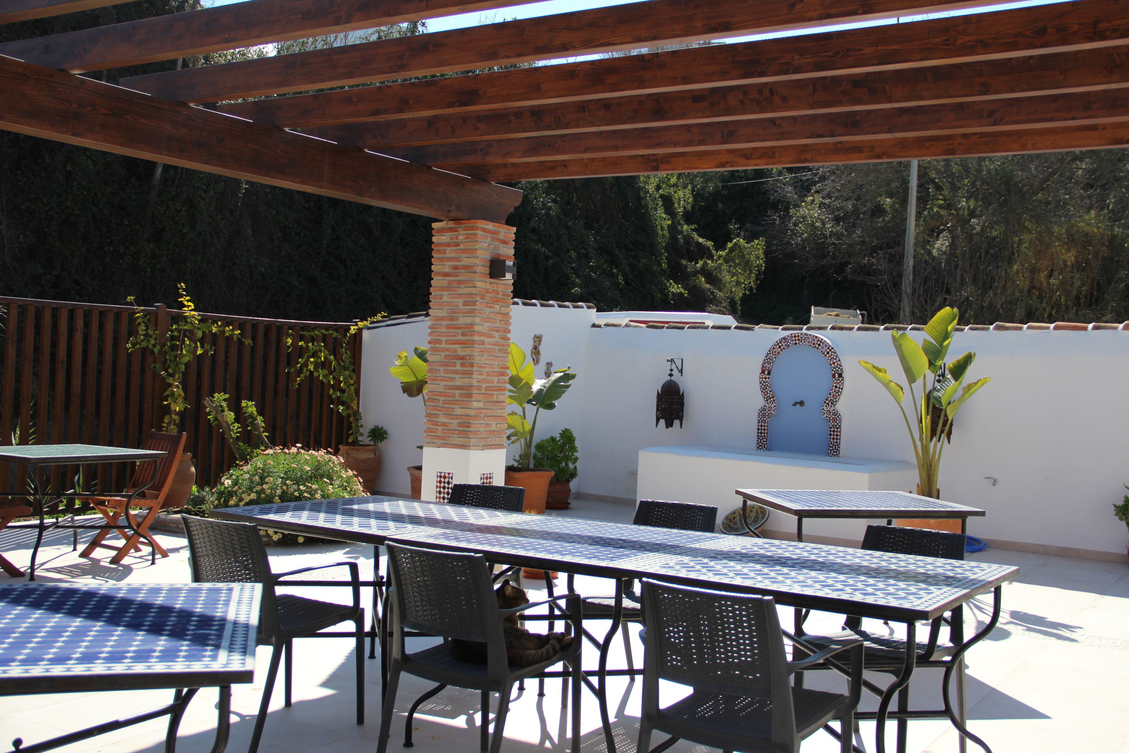 An outdoor patio with a table and chairs, surrounded by potted plants and a wooden pergola.
