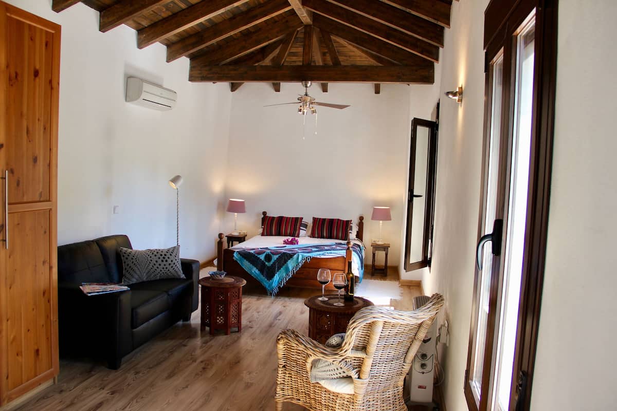A bedroom with a bed, a wooden ceiling, a ceiling fan, a window, a nightstand, a lamp, and a wicker chair.