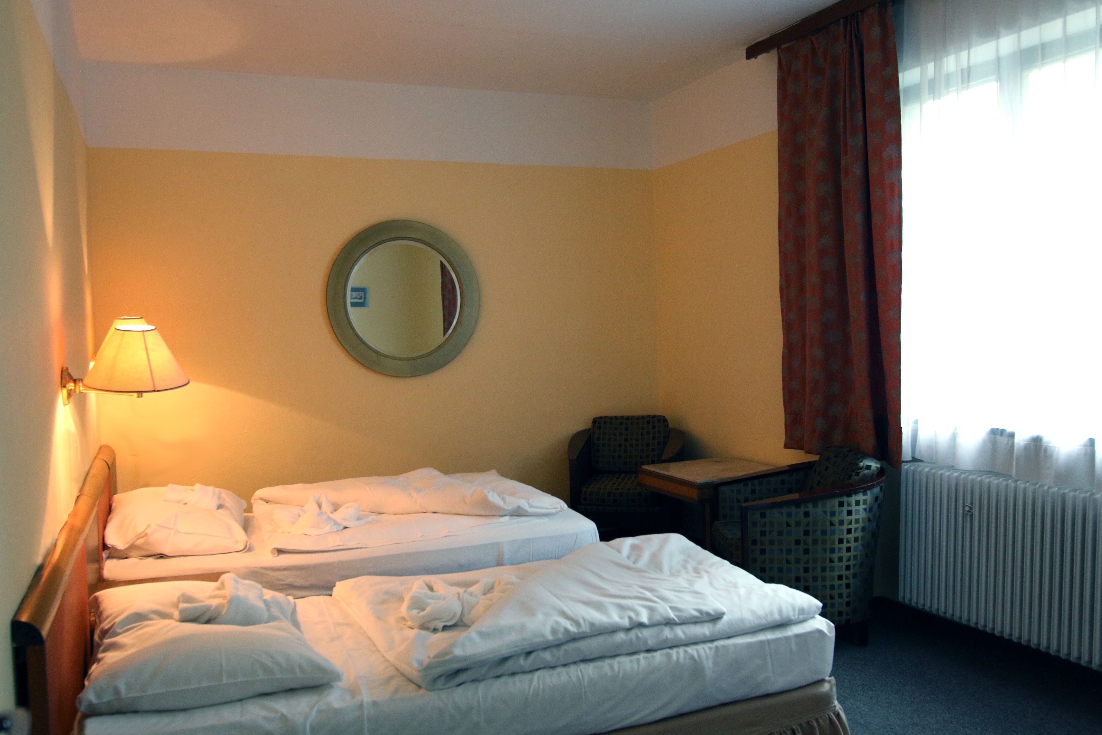 A hotel room with two beds, a round mirror, a chair, a table, and a lamp.