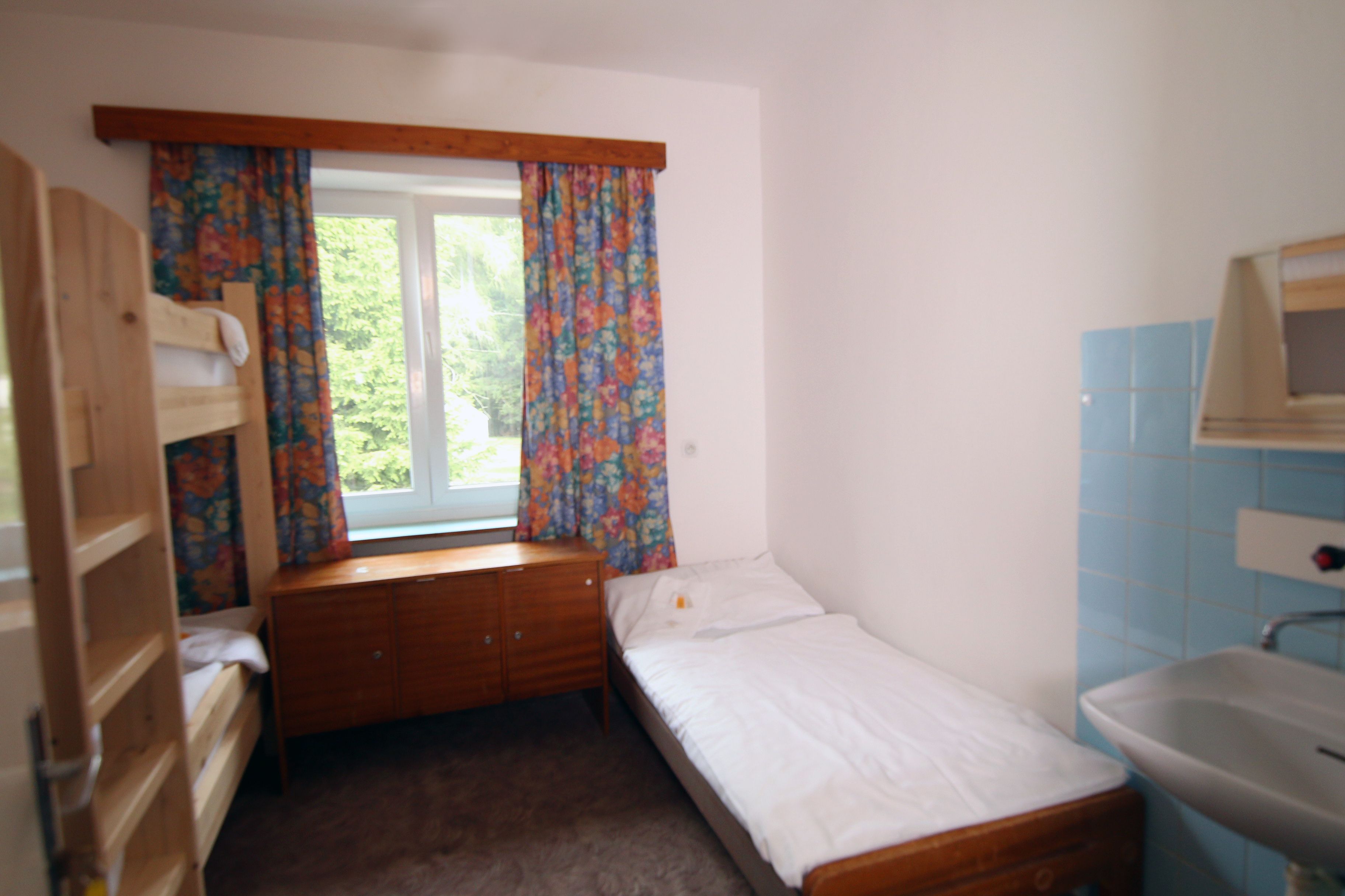 A small, single-bed room with a wooden bunk bed, a window with floral curtains, and a bathroom with a sink and mirror.