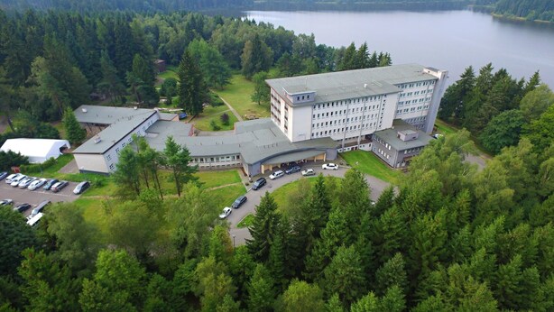 Vue aérienne d’un hôtel, avec un grand bâtiment, un parking et la forêt environnante.