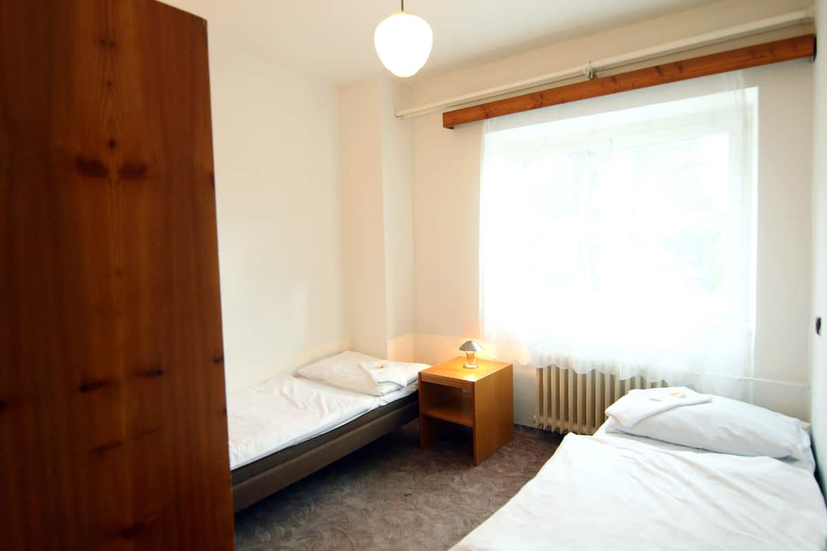 A small, clean room with a single bed, a bedside table with a lamp, a radiator, and a window with sheer curtains.