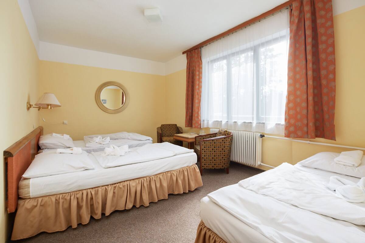 A hotel room with two beds, a round mirror, a table, a chair, and a window with curtains.