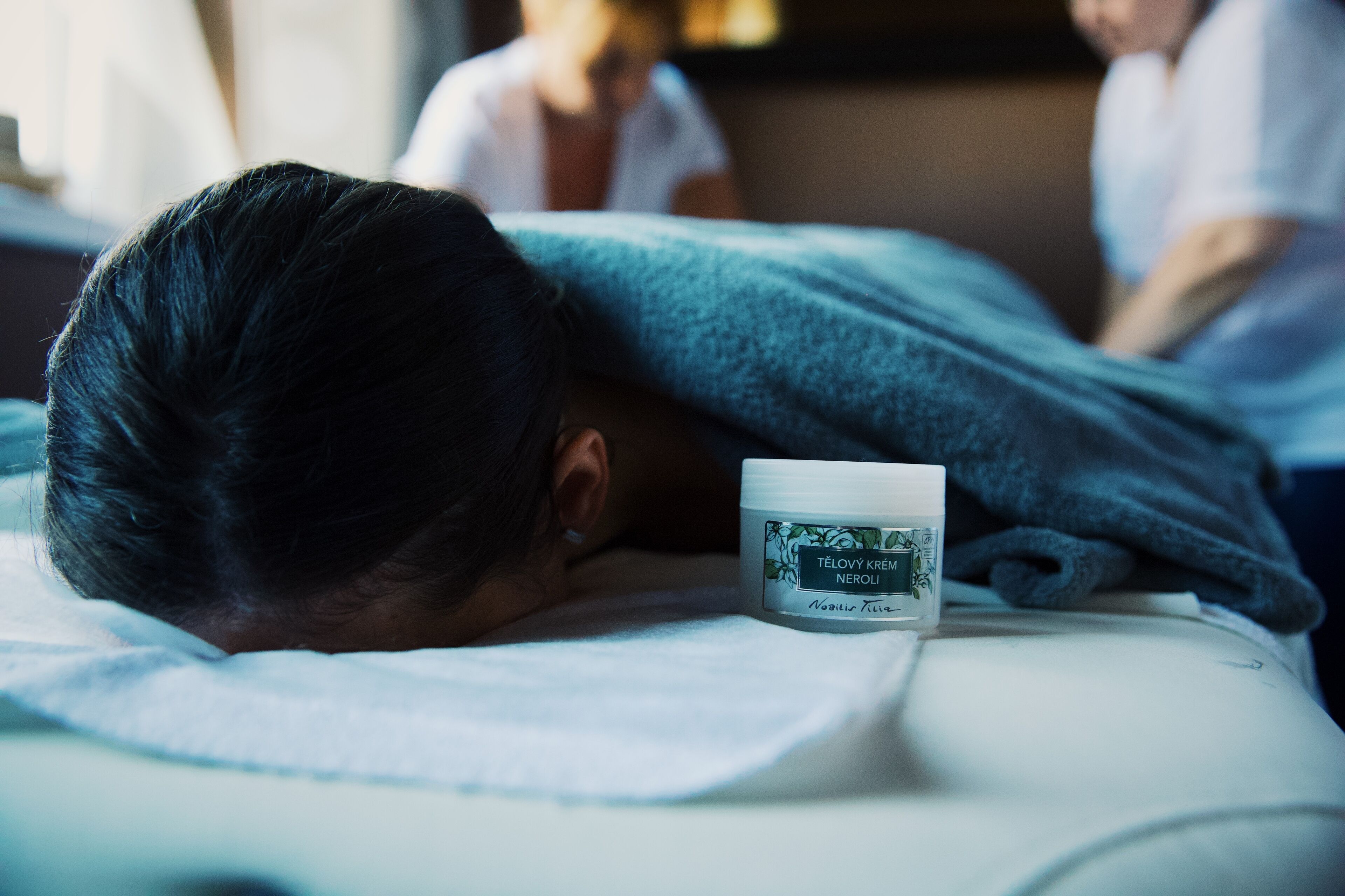 A person lying on a bed with a cream container labeled "TELOVY KREMLI NEROLI" and a blanket.