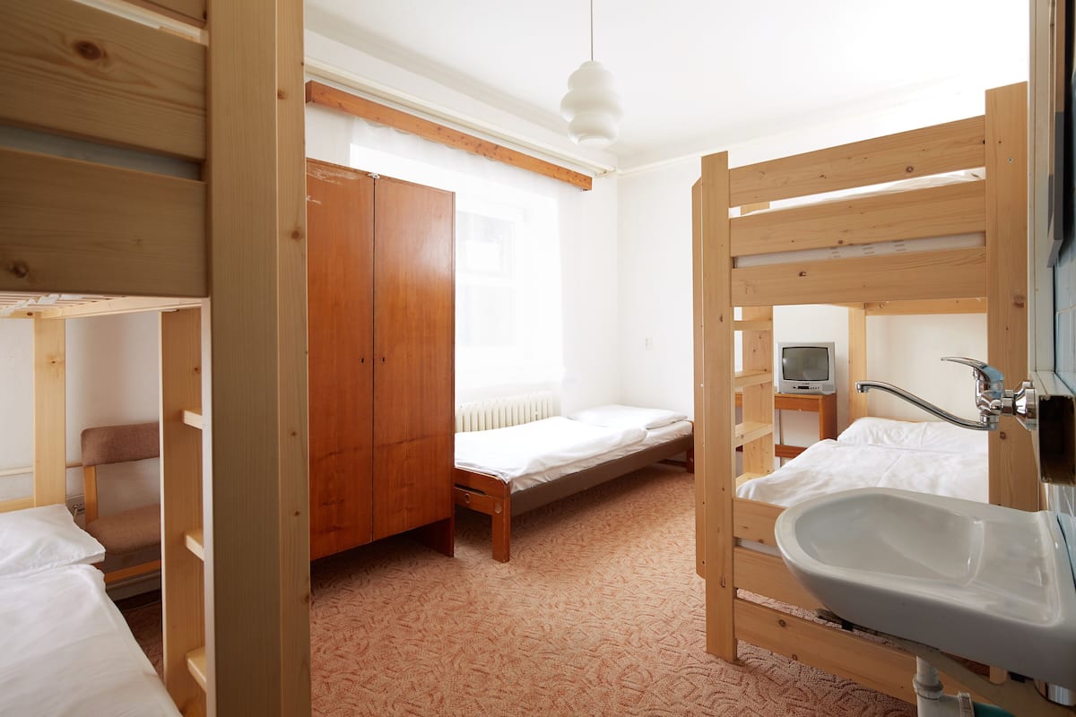 A room with bunk beds, a wooden wardrobe, a single bed, a bedside table with a television, and a bathroom with a sink and mirror.