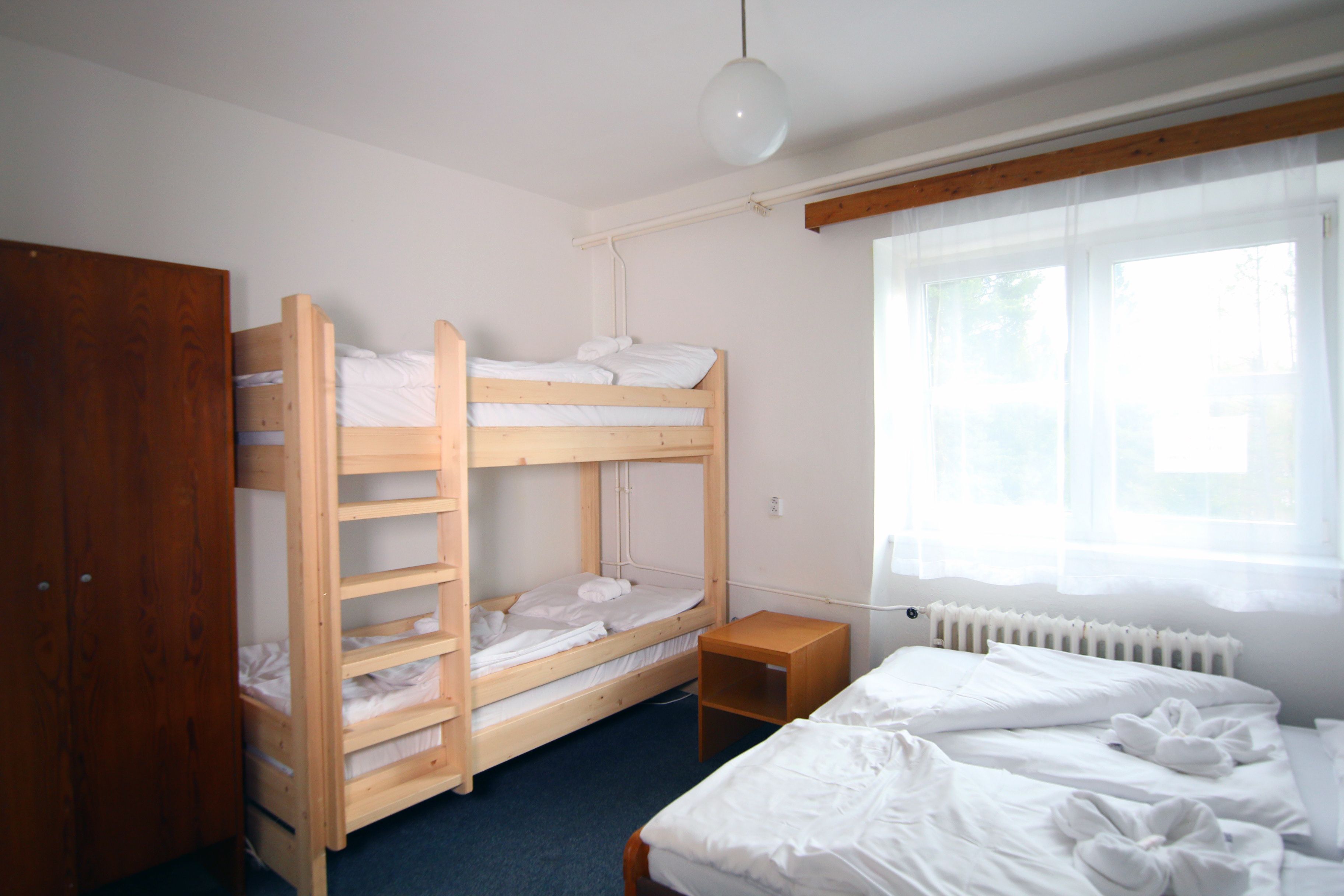 A bunk bed room with a wooden wardrobe, a small wooden nightstand, and a window with sheer curtains.