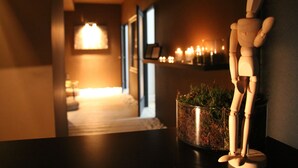 Un mannequin en bois est posé sur une table, à côté se trouve un pot de fleurs, une bougie allumée est posée sur une étagère, et un couloir est visible en arrière-plan, avec une lampe et des bougies allumées.