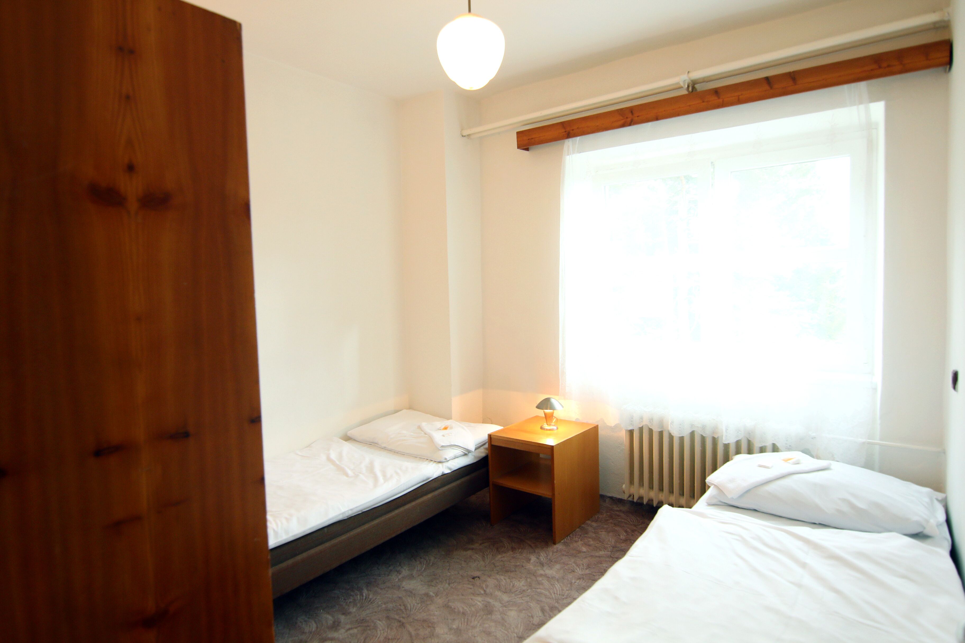 A small, tidy room with a single bed, a nightstand with a lamp, and a window with sheer curtains.