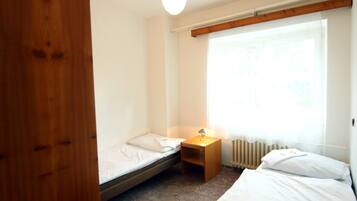 A small, tidy room with a single bed, a nightstand with a lamp, and a window with sheer curtains.