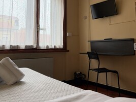 Una camera d'albergo con un letto, una sedia, una TV a parete e una finestra con tende leggere.