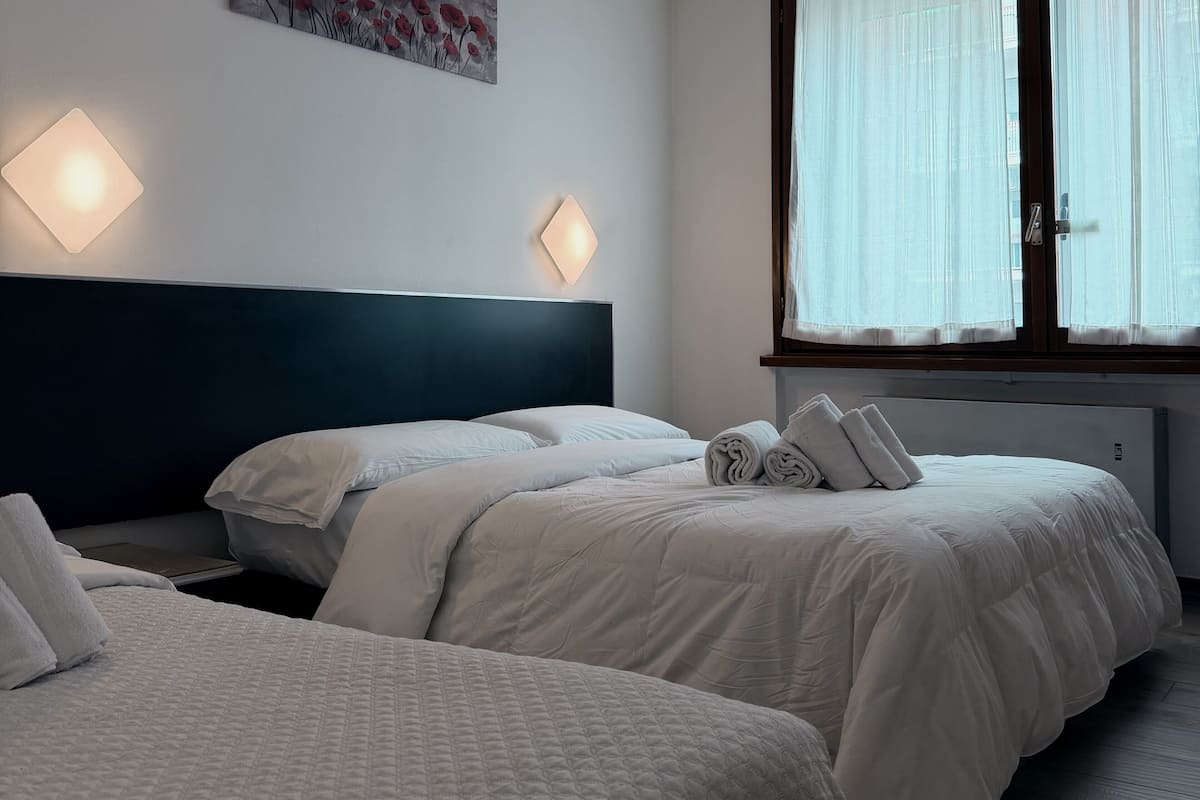 Habitación de hotel con dos camas, un cabecero oscuro y una ventana con cortinas traslúcidas.
