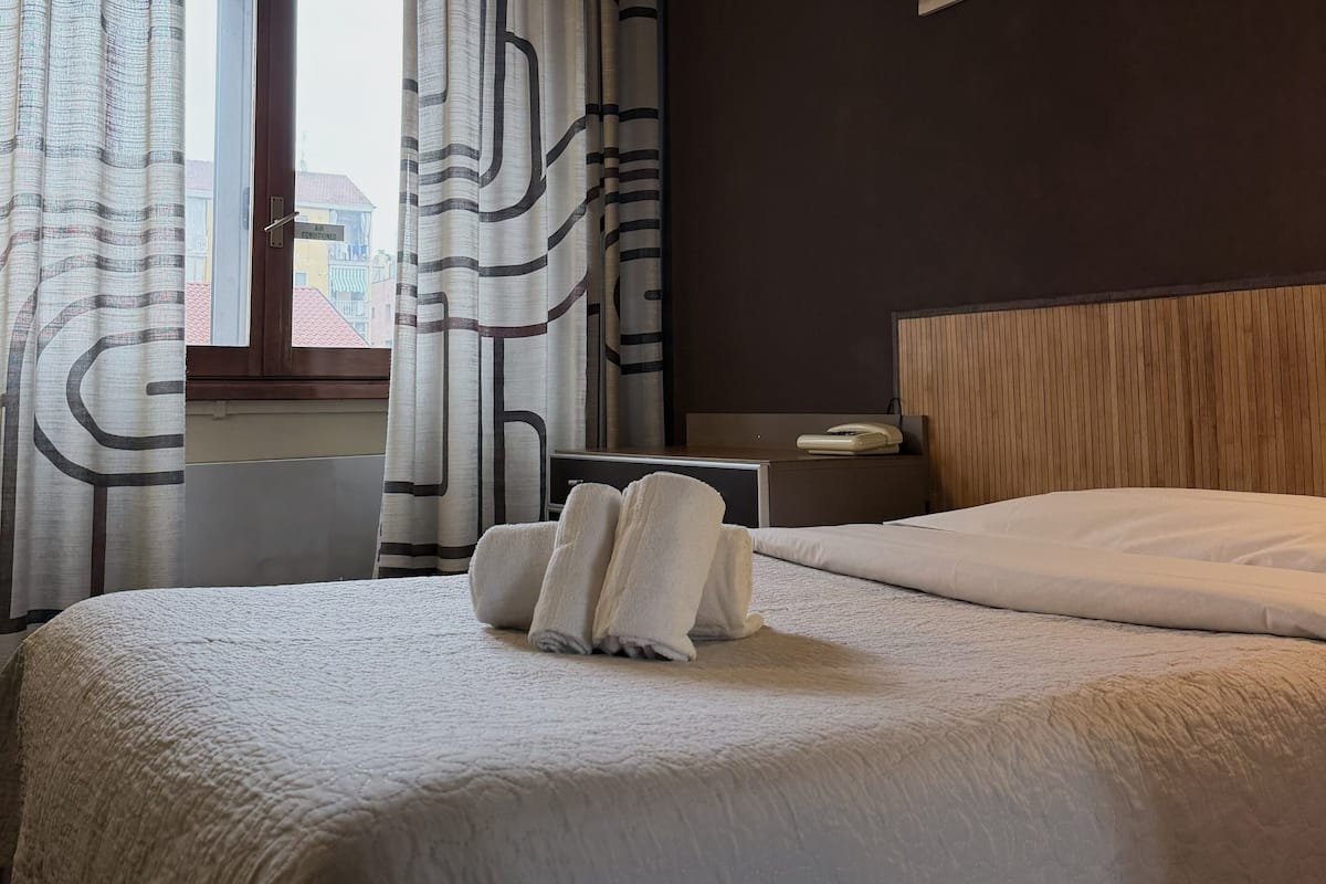 Habitación de hotel con cama, mesita de noche, teléfono y ventana con cortinas.