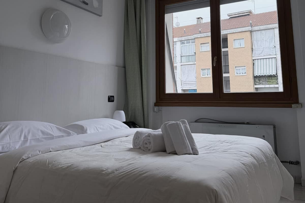 Habitación de hotel con una cama grande, una mesita de noche, una lámpara y una ventana con vista a edificios.