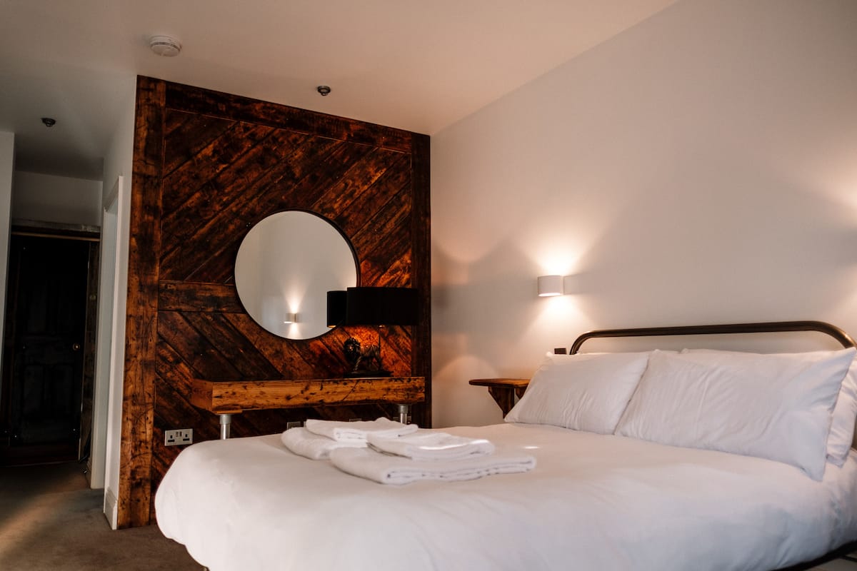 A hotel room with a large bed, a wooden accent wall, a round mirror, and a nightstand with a lamp.