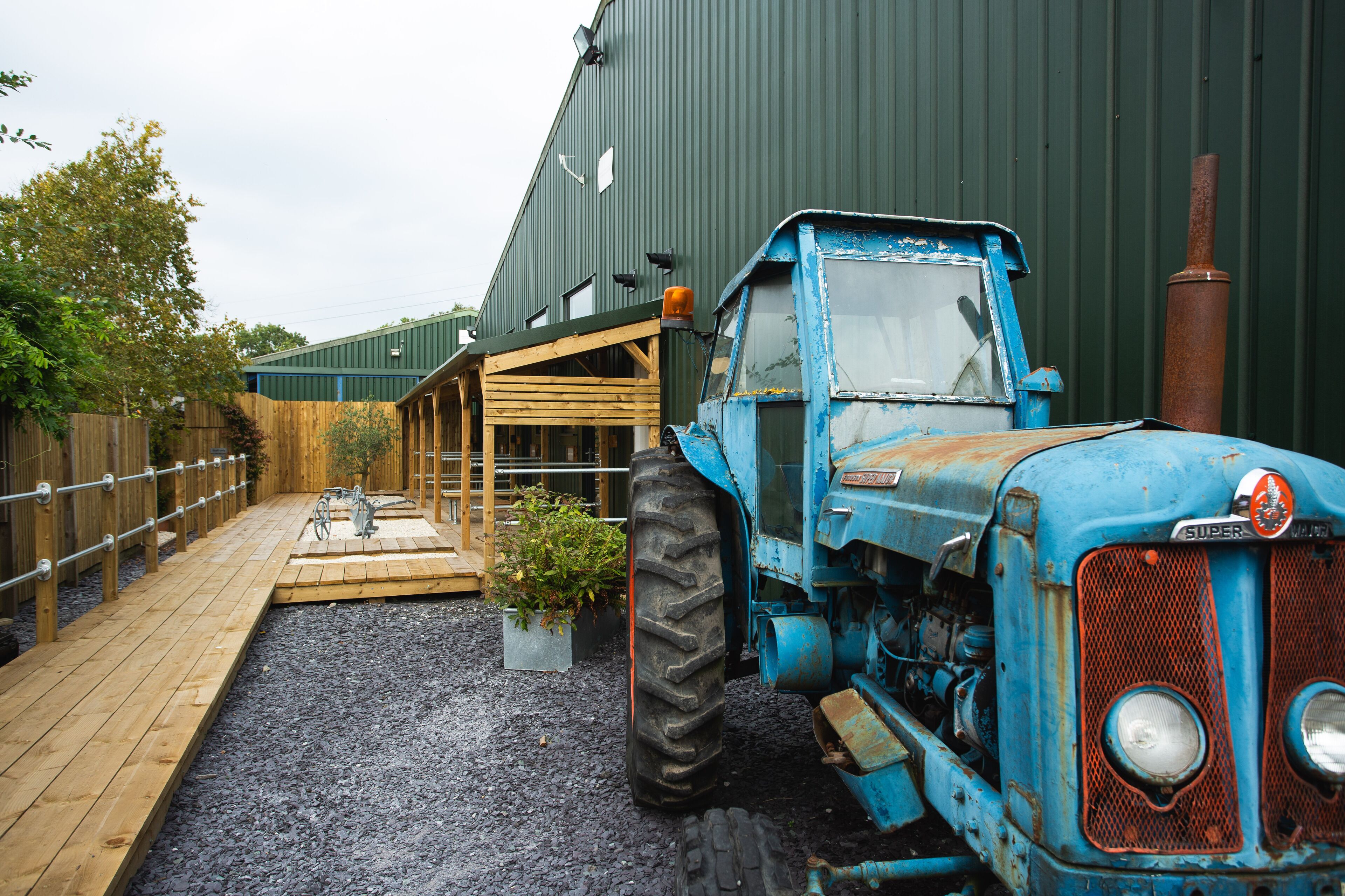 Un tractor azul estacionado frente a un edificio verde con una terraza de madera y una planta en maceta.