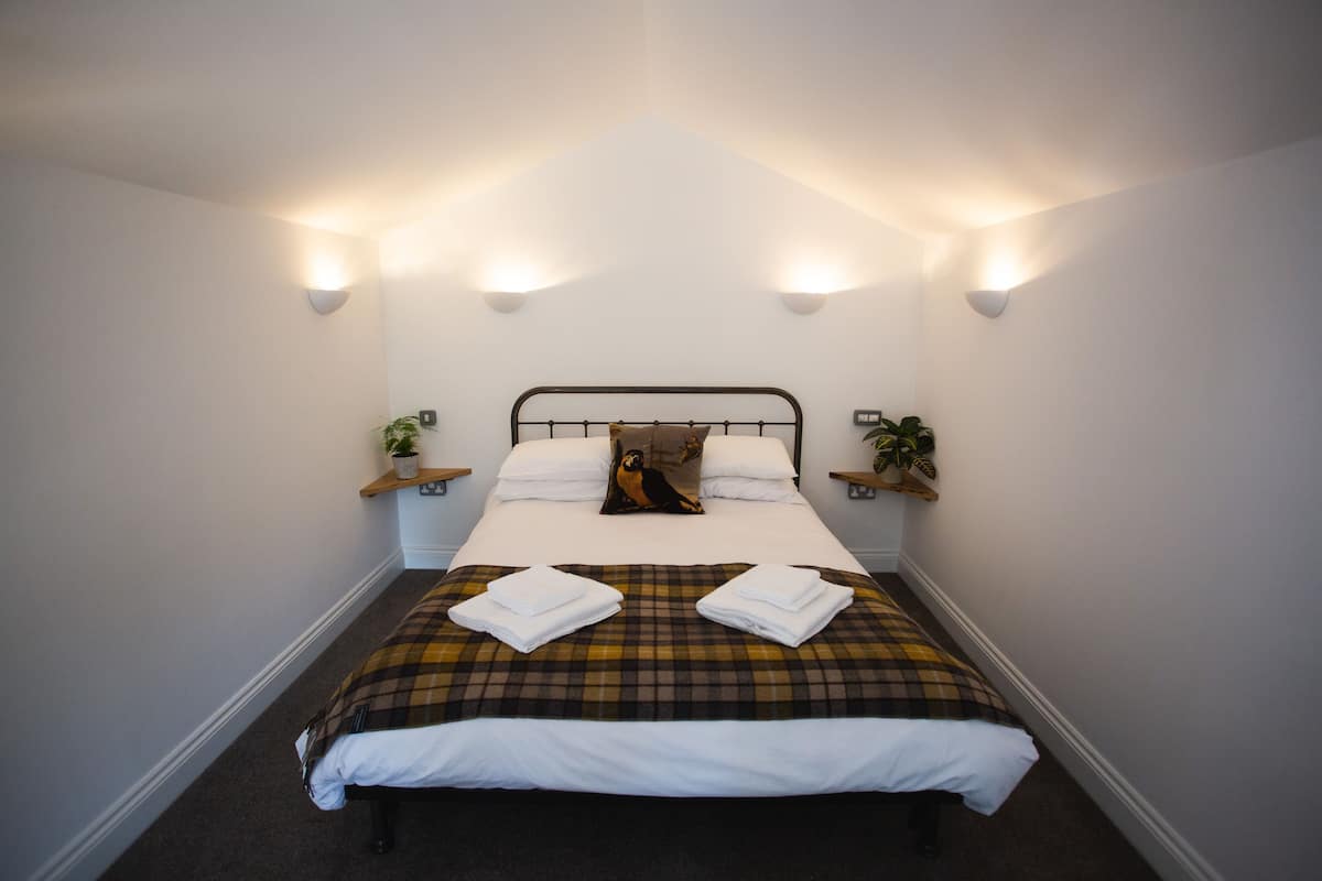 A neatly made bed with a plaid bedspread, two pillows, and a decorative pillow. There are two bedside tables with plants and wall-mounted lights.