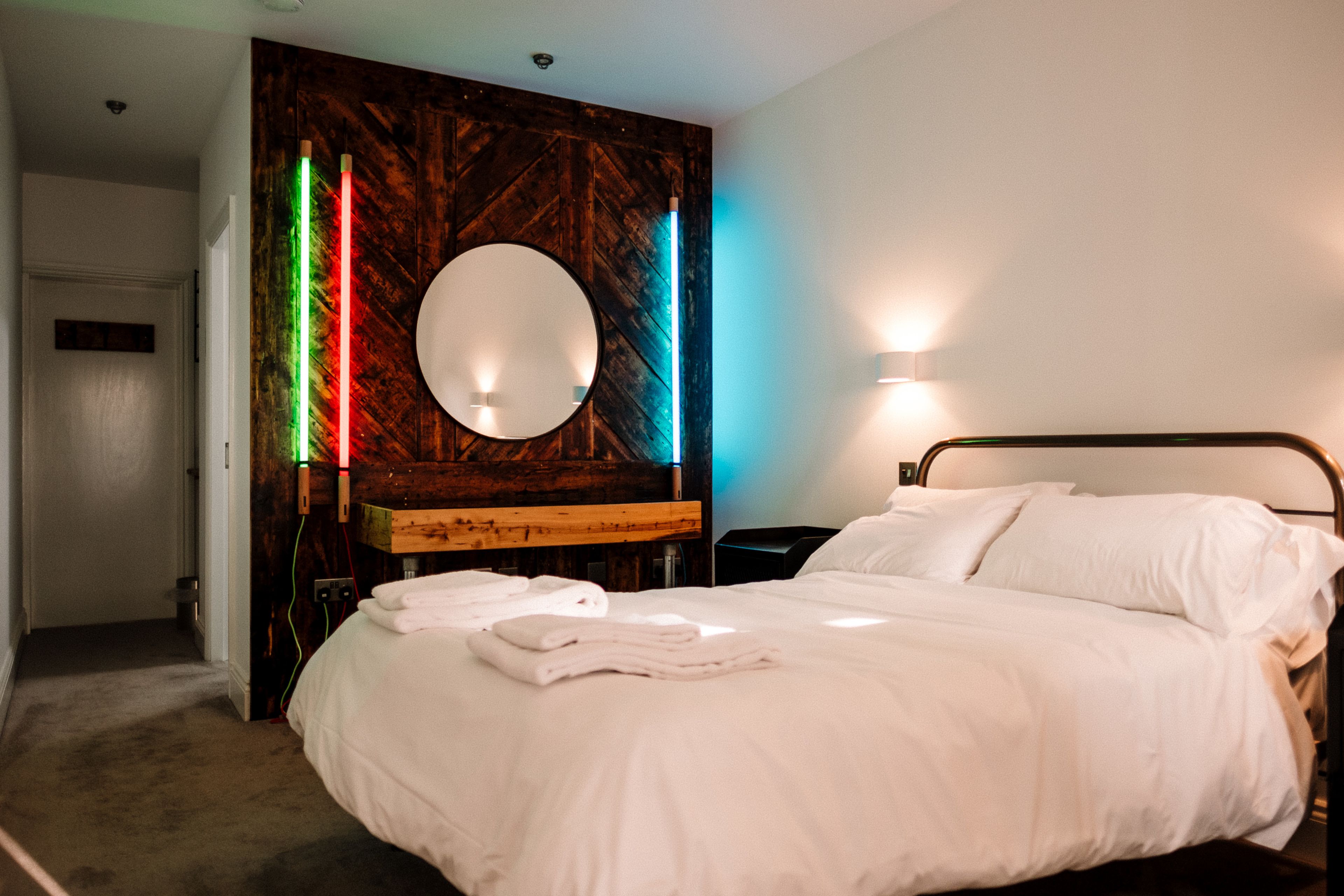 Habitación de hotel con una cama grande, un espejo redondo, un panel de madera decorativo y una mesita de noche con lámpara.