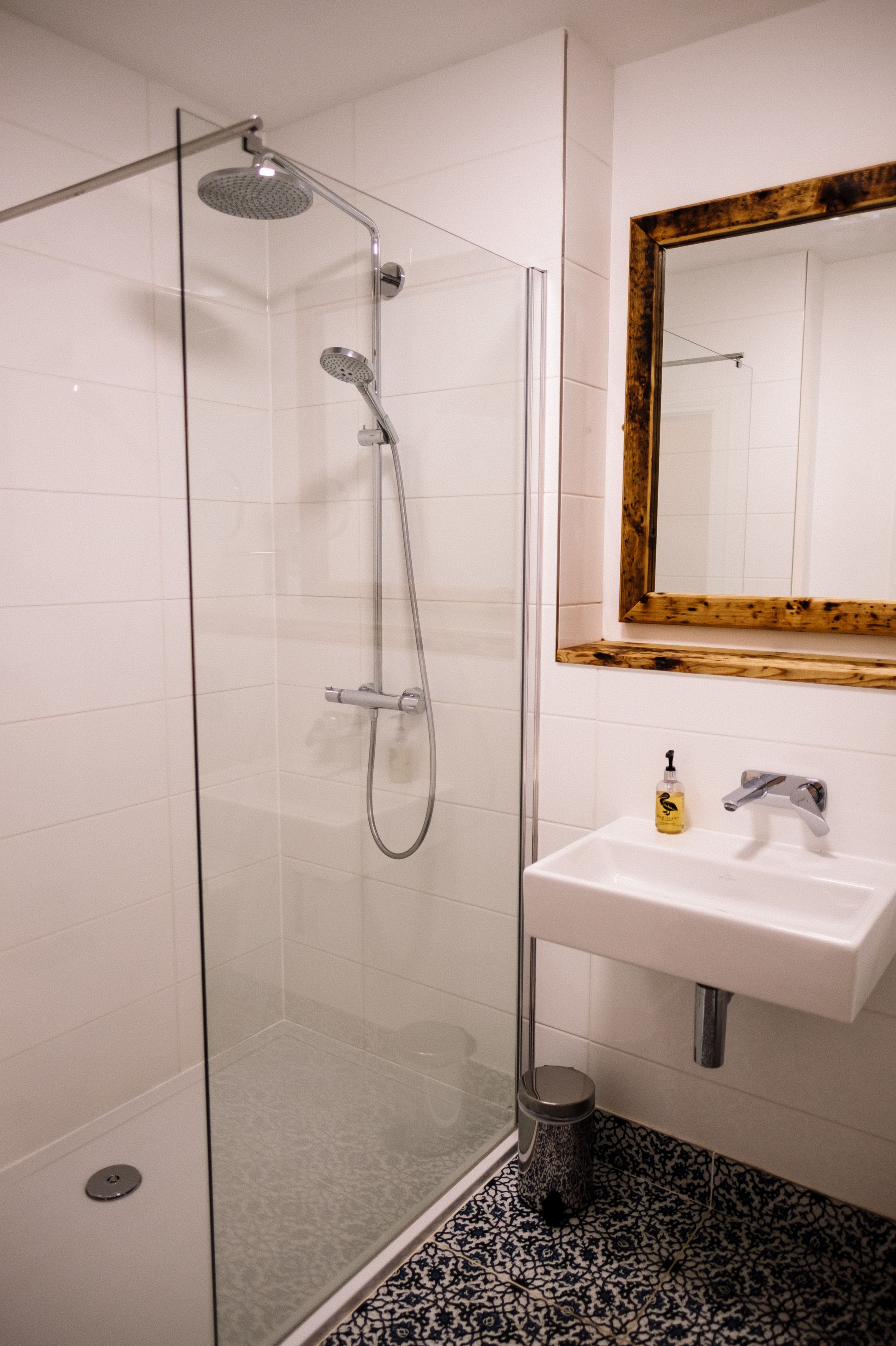 A glass-enclosed shower with a handheld showerhead and a wall-mounted faucet. Adjacent to the shower is a white sink with a modern faucet and a soap dispenser. A trash can is placed next to the sink.