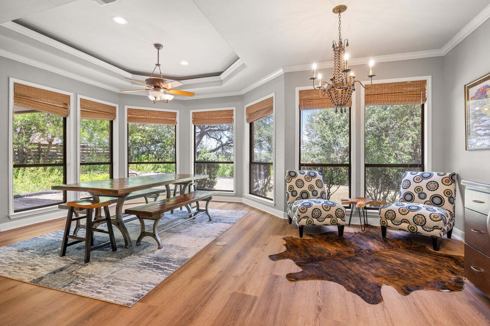 A spacious room with a dining table, benches, patterned chairs, a chandelier, large windows with blinds, and a wooden floor.