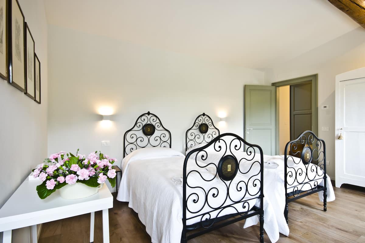 A bedroom with two beds, a white table with a vase of flowers, and a door leading to another room.