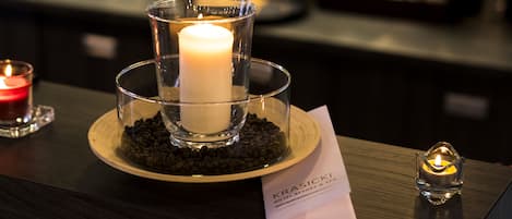 Una vela en un portavelas de vidrio sobre una bandeja de madera, con otra vela encendida al fondo y un portavelas con vela encendida a la derecha.