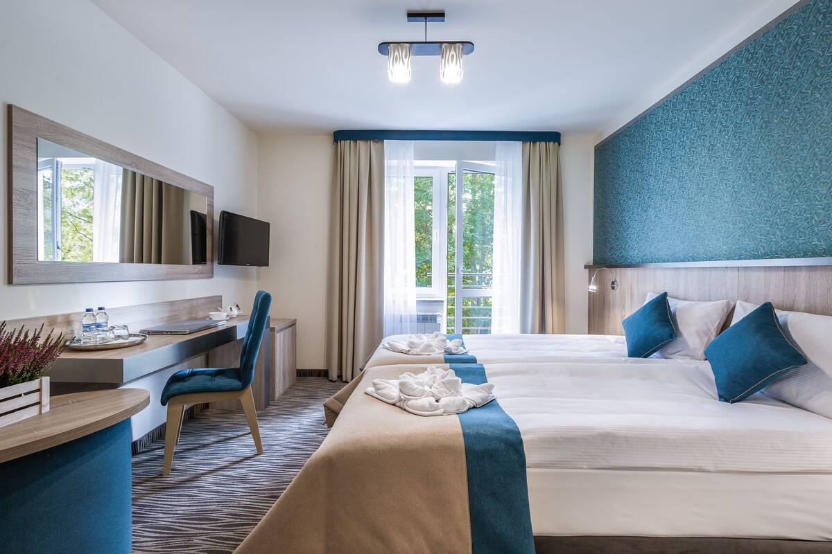 A hotel room with two beds, a desk, a chair, a TV, and a large window with curtains.