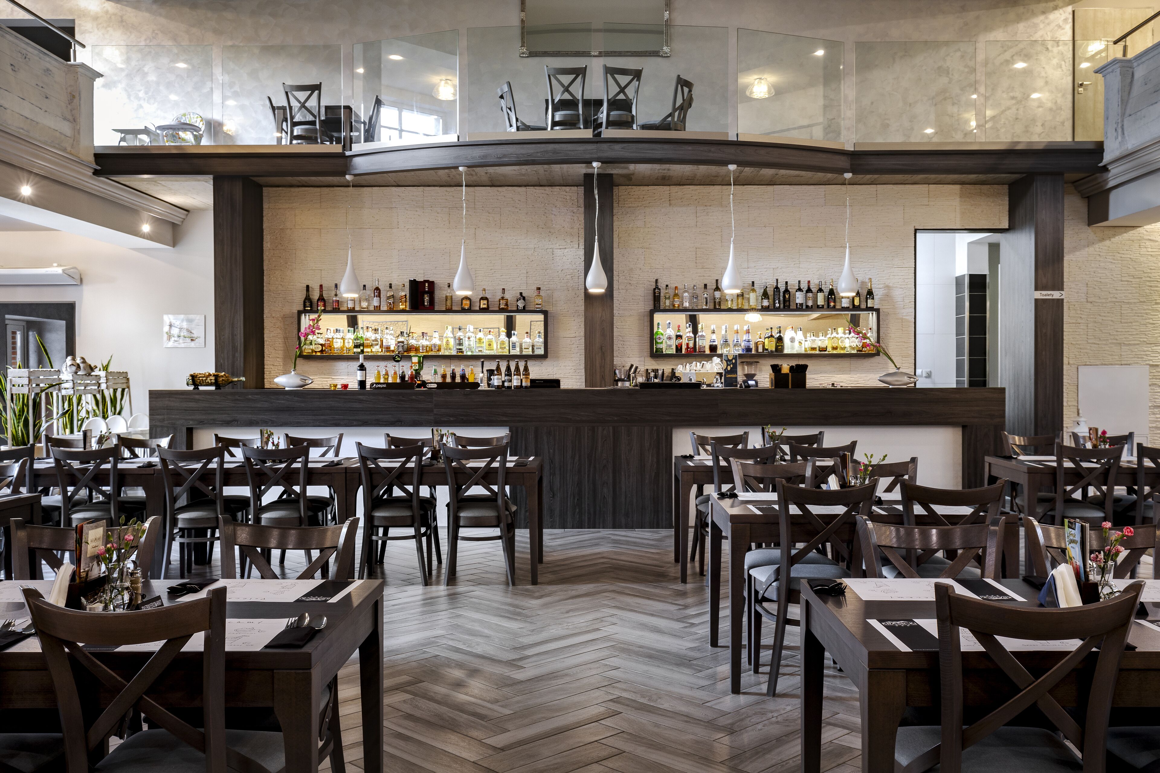 A restaurant with a herringbone floor, wooden chairs, and a bar with bottles.