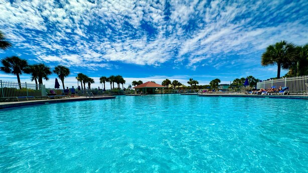 Une piscine d’un bleu éclatant, avec des chaises longues et des palmiers, sous un ciel partiellement nuageux.