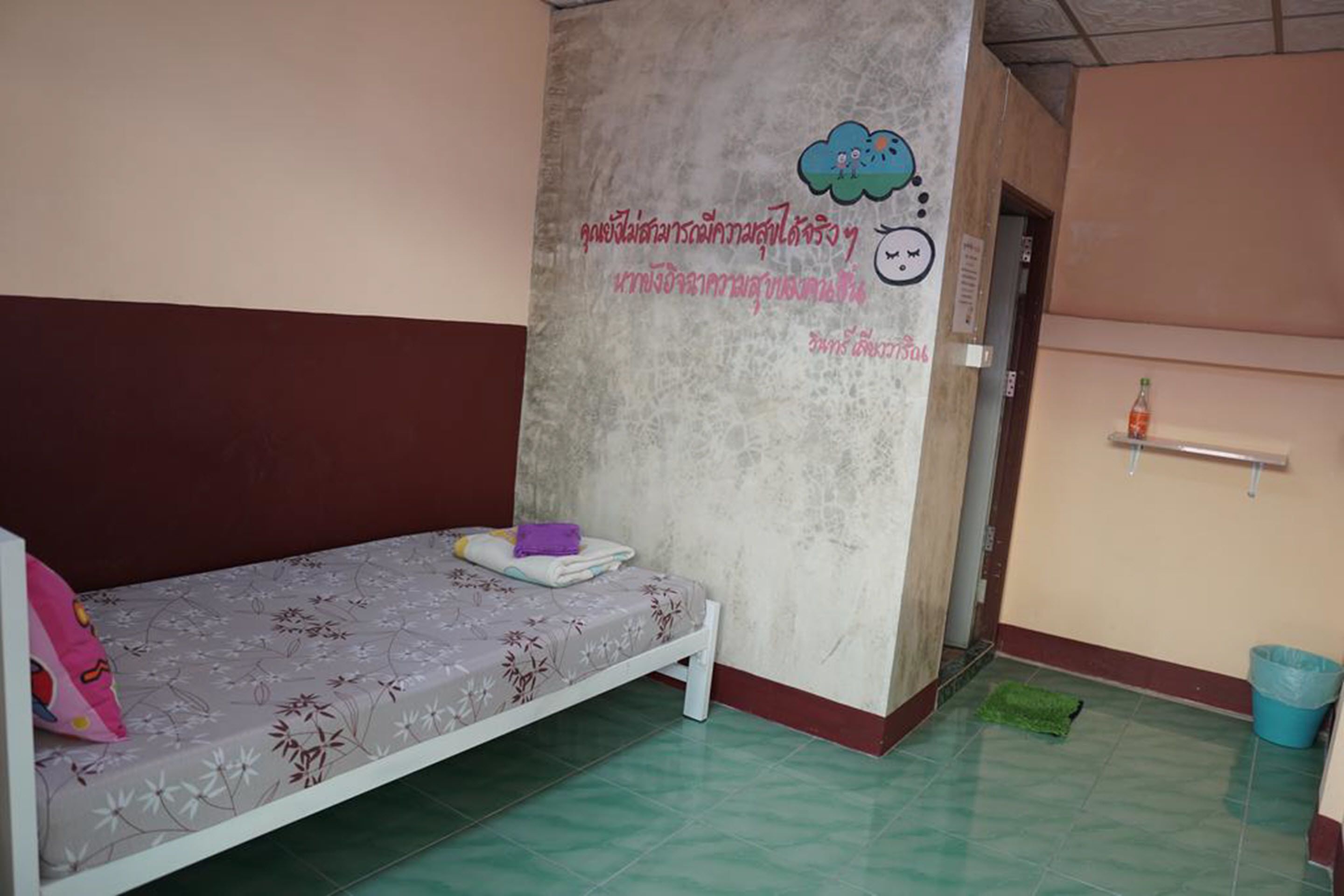 A single-bed room with a patterned bedspread, a wall with a decorative design, a trash bin, and a small shelf with a bottle.