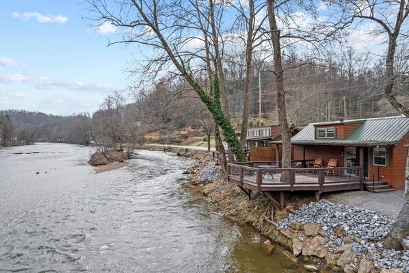 Une cabane au bord d’une rivière, dotée d’une terrasse en bois et d’un espace salon extérieur.