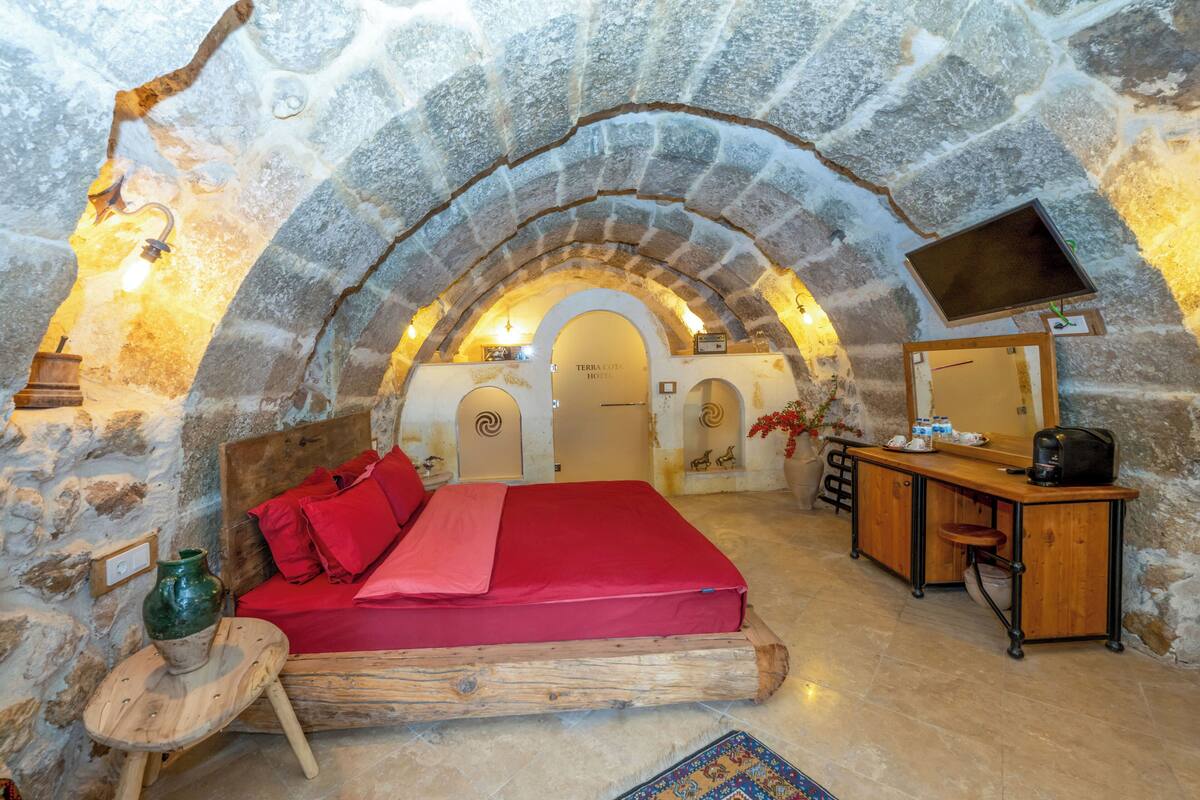 A cozy room with a stone arch ceiling, a bed with red bedding, a wooden desk, and a television.