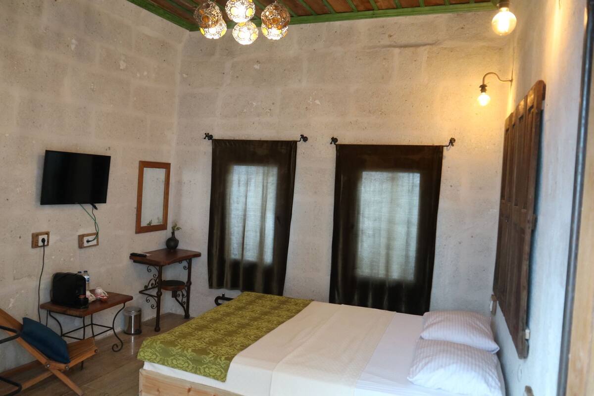 A room with a bed, a desk, a chair, a television, and two windows with curtains.