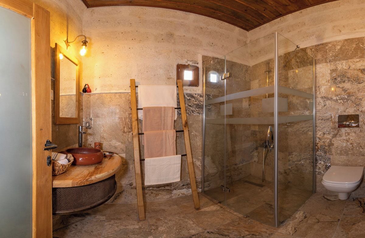 A rustic bathroom with a stone wall, a wooden ladder, a glass shower enclosure, and a round sink with a basket.