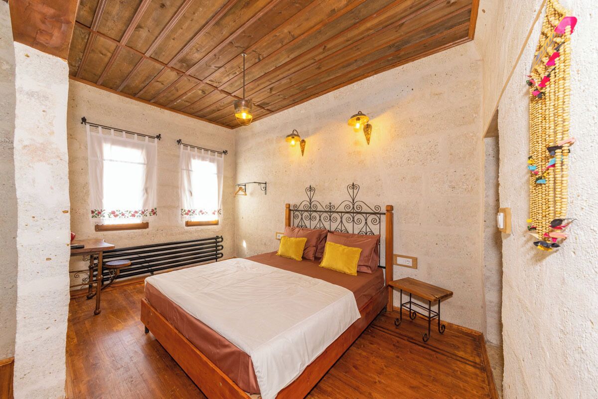 A bedroom with a wooden ceiling, a bed with a metal headboard, two wall-mounted lights, a small wooden table, and a window with curtains.