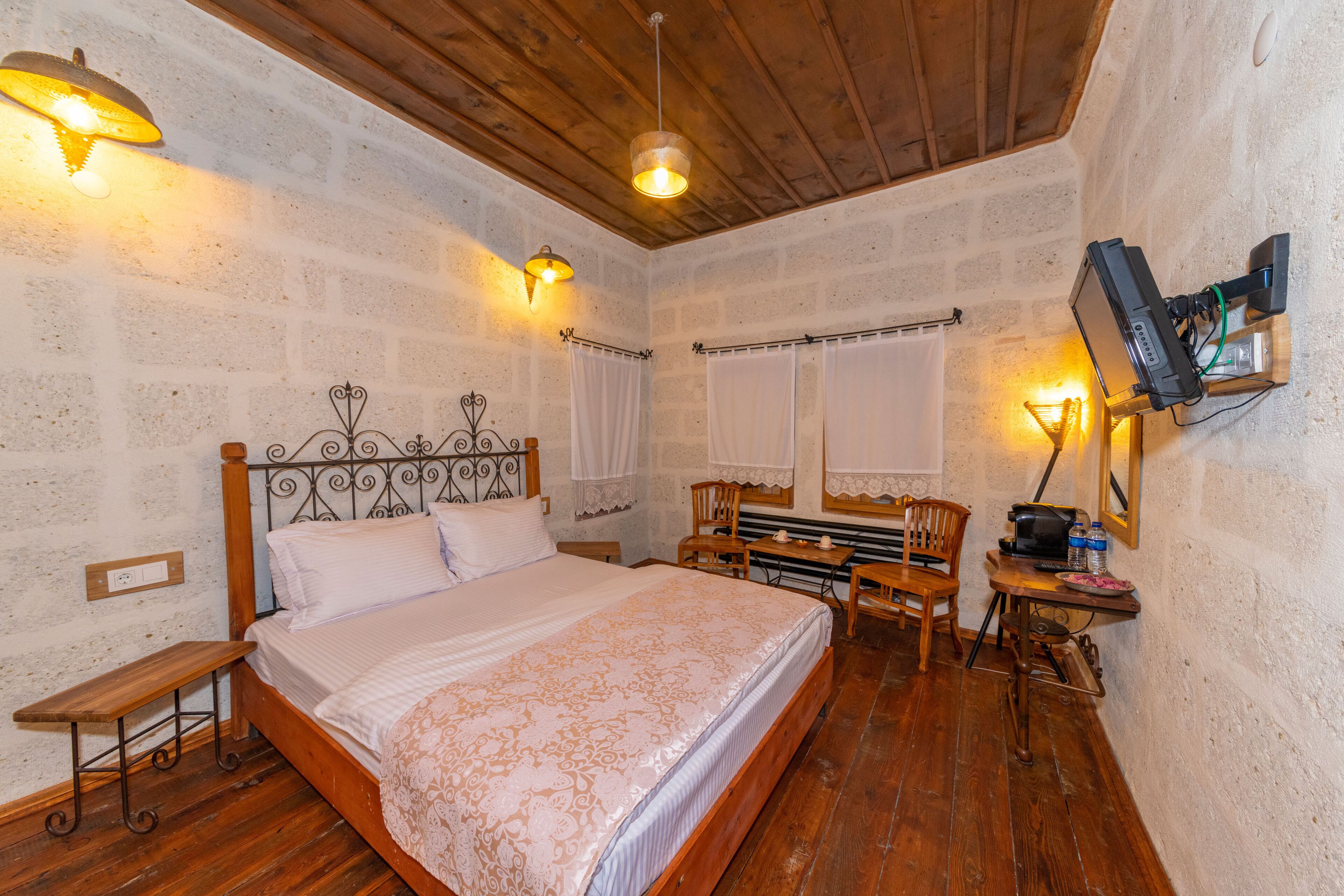 A bedroom with a wooden bed, a desk with a chair, a television mounted on the wall, and a window with curtains.