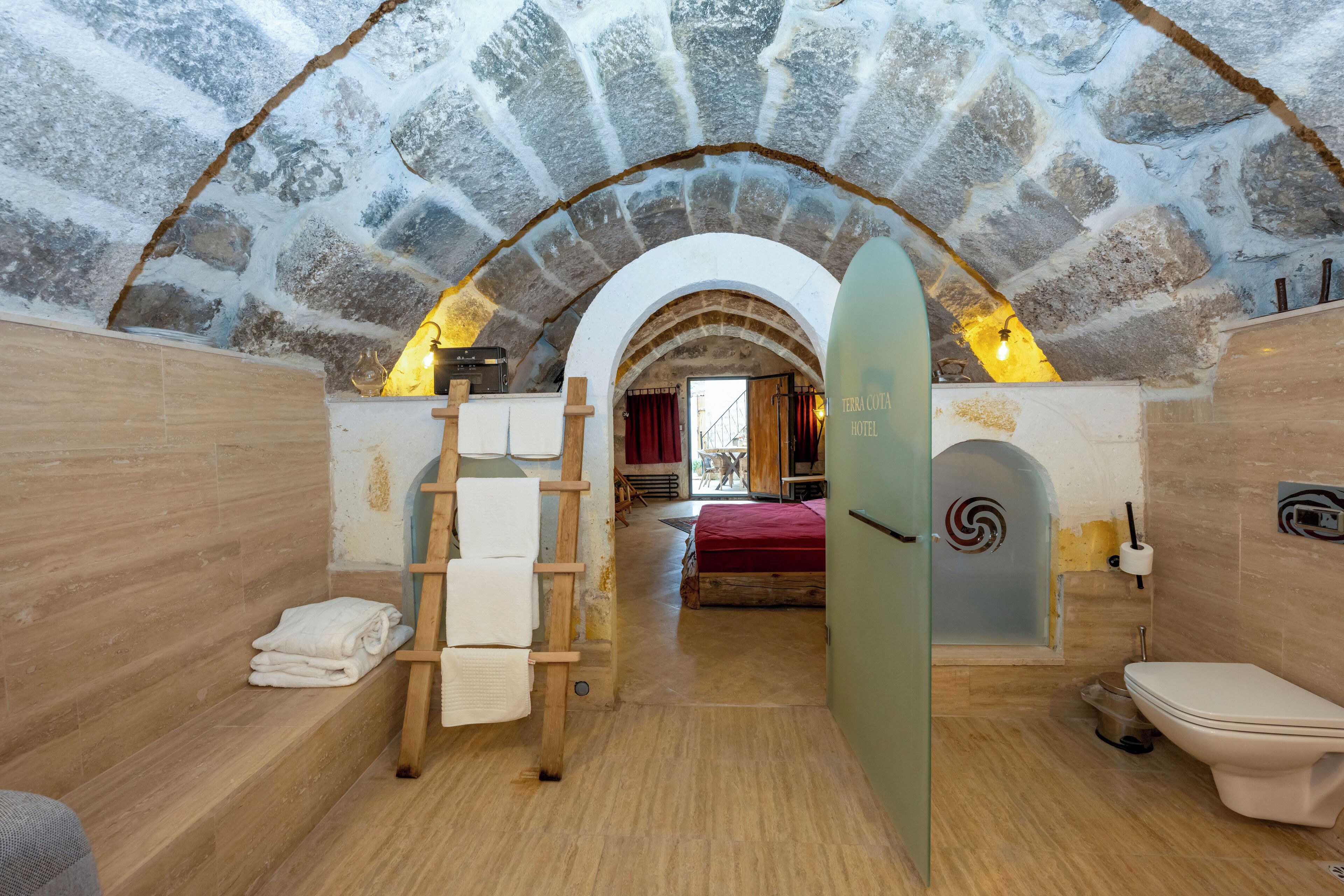 A stone-walled bathroom with a wooden ladder, a red bed, and a glass door leading to a room.