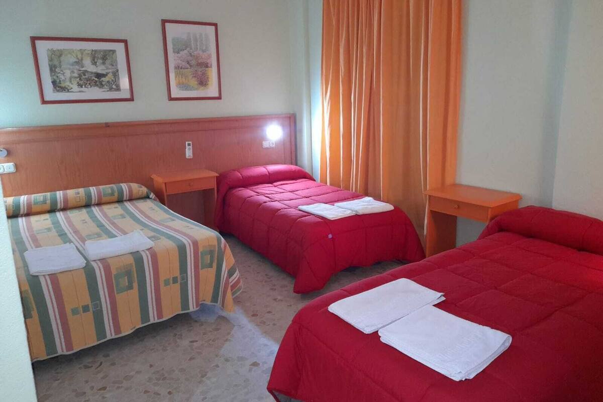 A room with two beds, one with a striped bedspread and the other with a solid red bedspread. There are two bedside tables, one with a lamp, and two framed pictures on the wall.