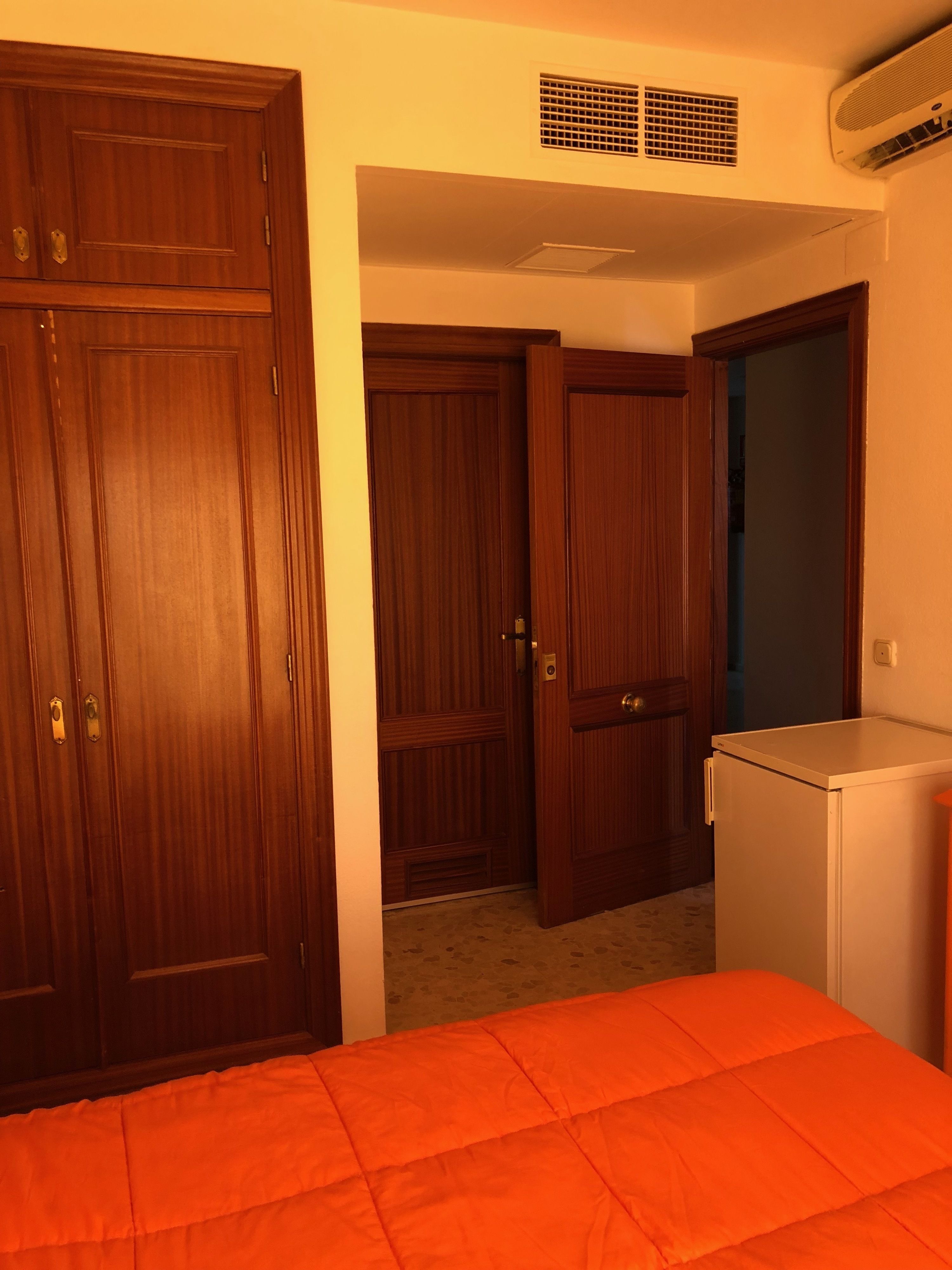 A bedroom with a bed, a wardrobe, a small refrigerator, and a door leading to another room.