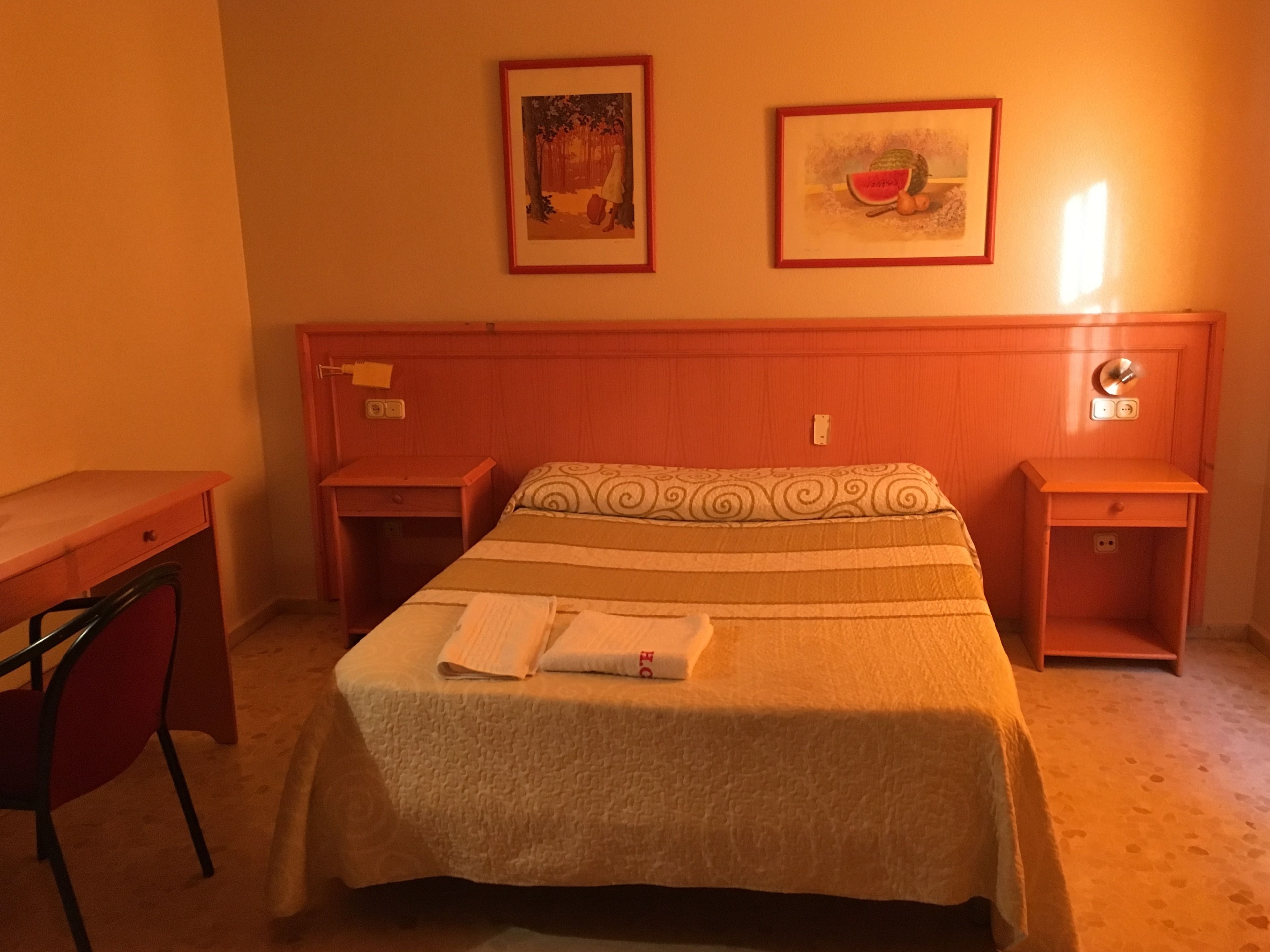 A hotel room with a bed, two nightstands, a desk, and two framed pictures on the wall.