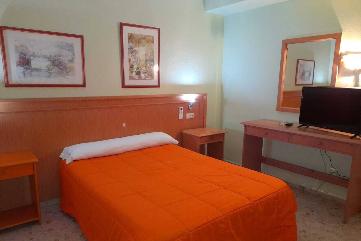A hotel room with a bed, a desk, a television, and two framed pictures on the wall.