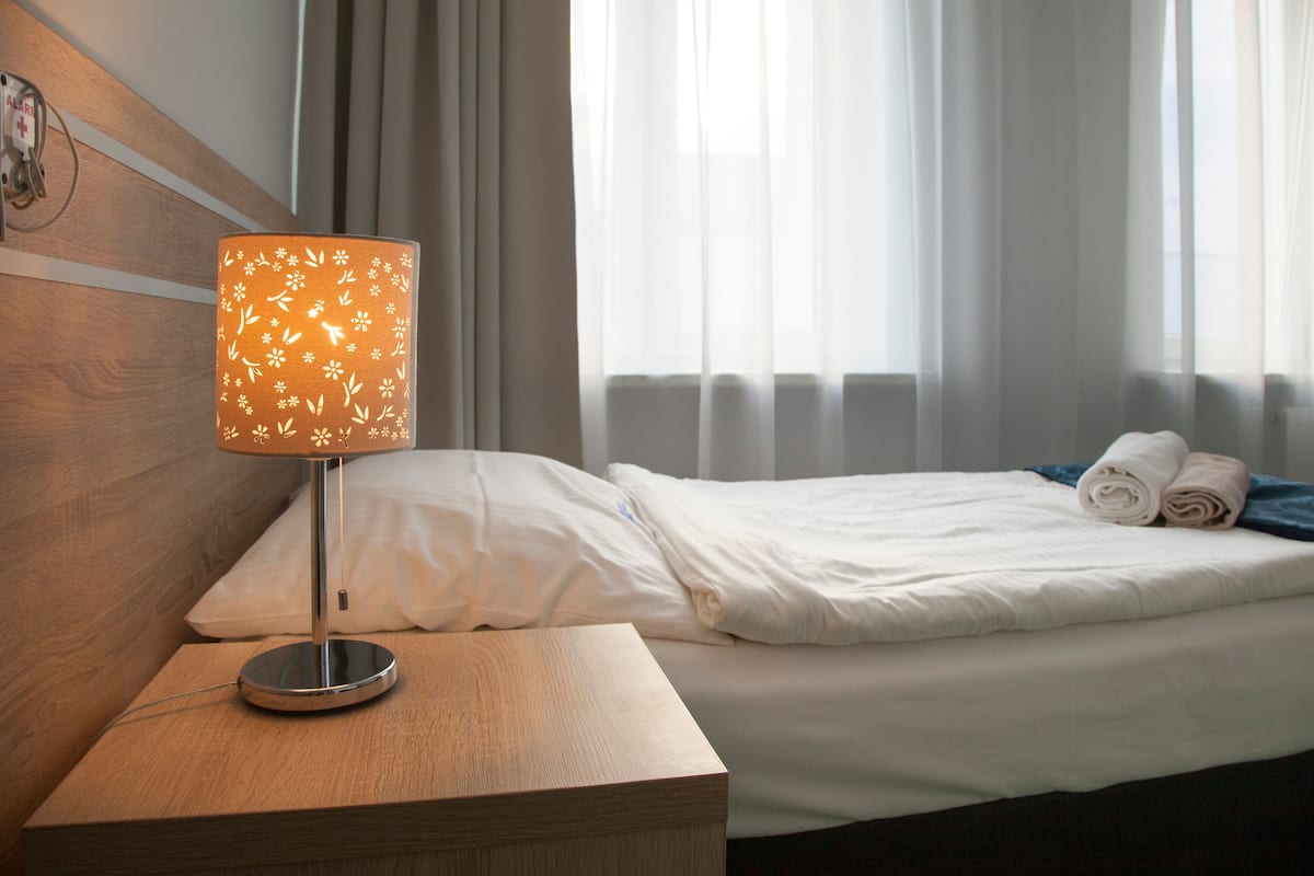 A hotel room with a bed, a bedside table with a lamp, and a window with curtains.