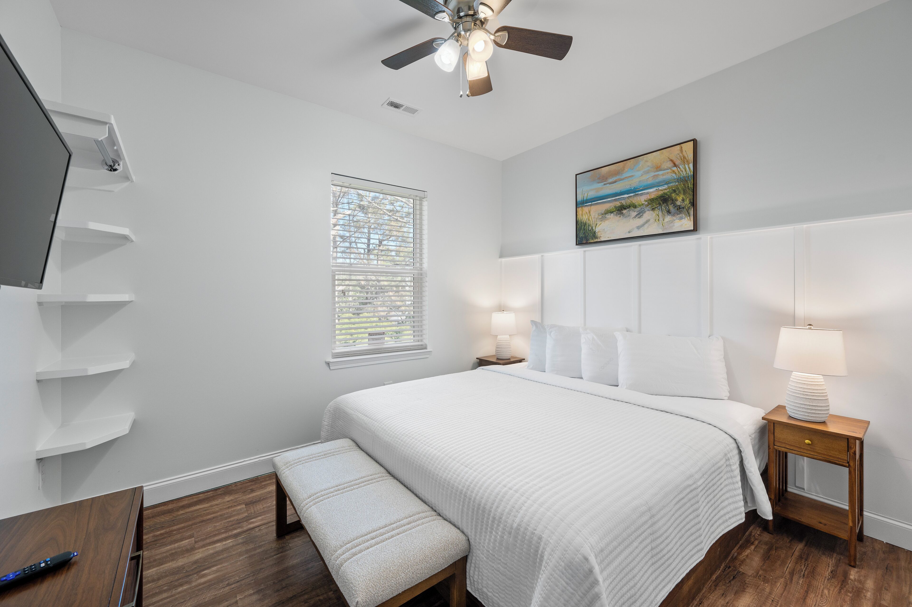 A bedroom with a bed, a bench, a TV, a ceiling fan, a window, and a painting on the wall.
