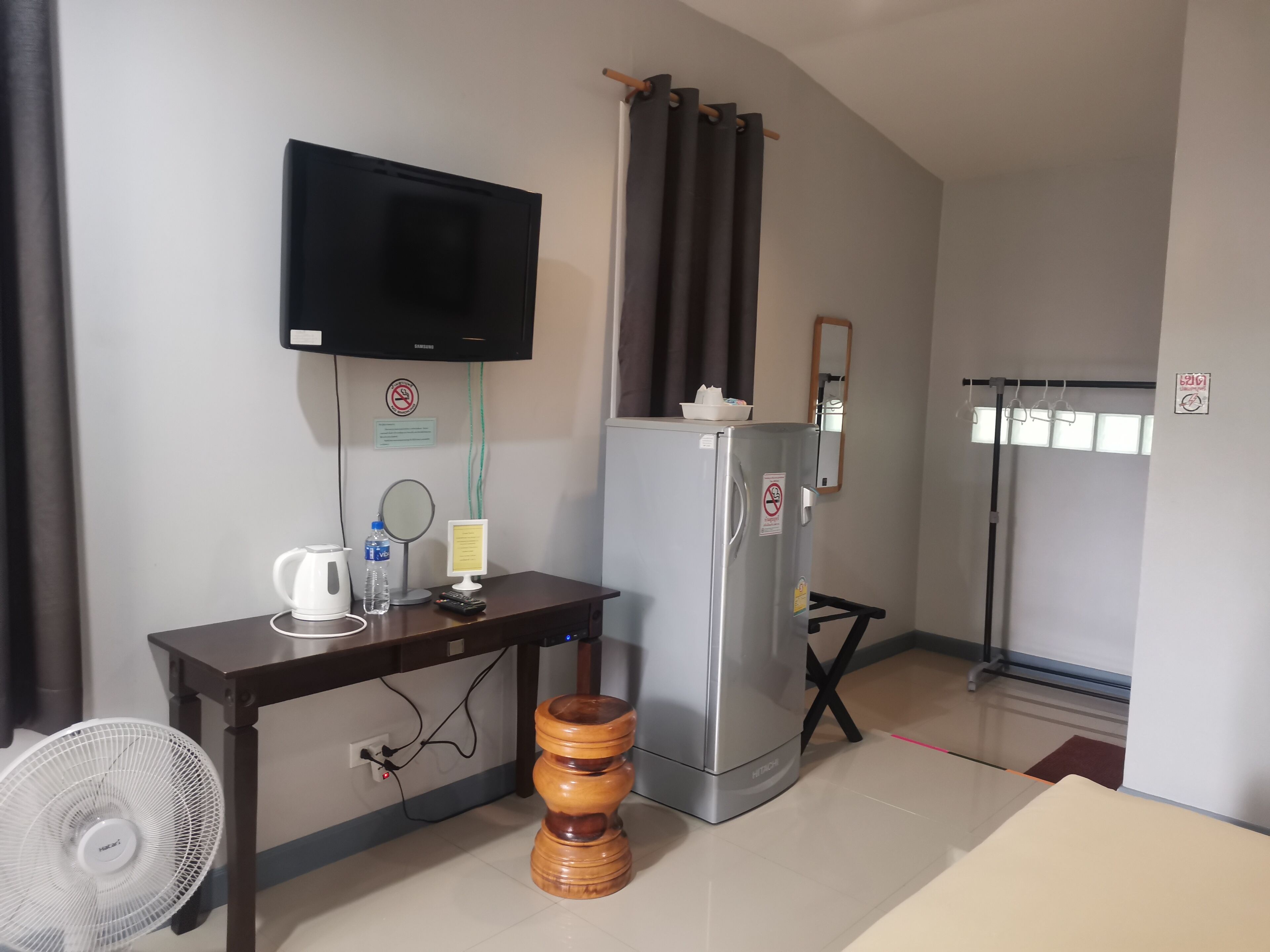 Habitación de hotel con televisión de pantalla plana en la pared, refrigerador, taburete de madera, mesa con tetera, botella de agua y un espejo pequeño.