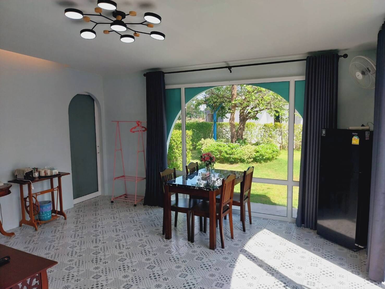 A dining area with a table and chairs, a hanging clothes rack, a ceiling light fixture, and a view of a garden through a large window.