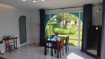 A dining area with a table and chairs, a hanging clothes rack, a ceiling light fixture, and a view of a garden through a large window.