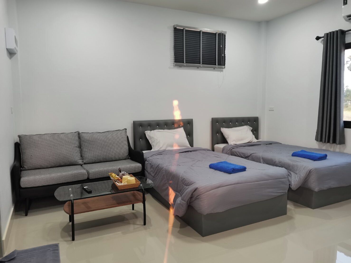 A room with a sofa, two beds, a small table with a basket, and a window with blinds.